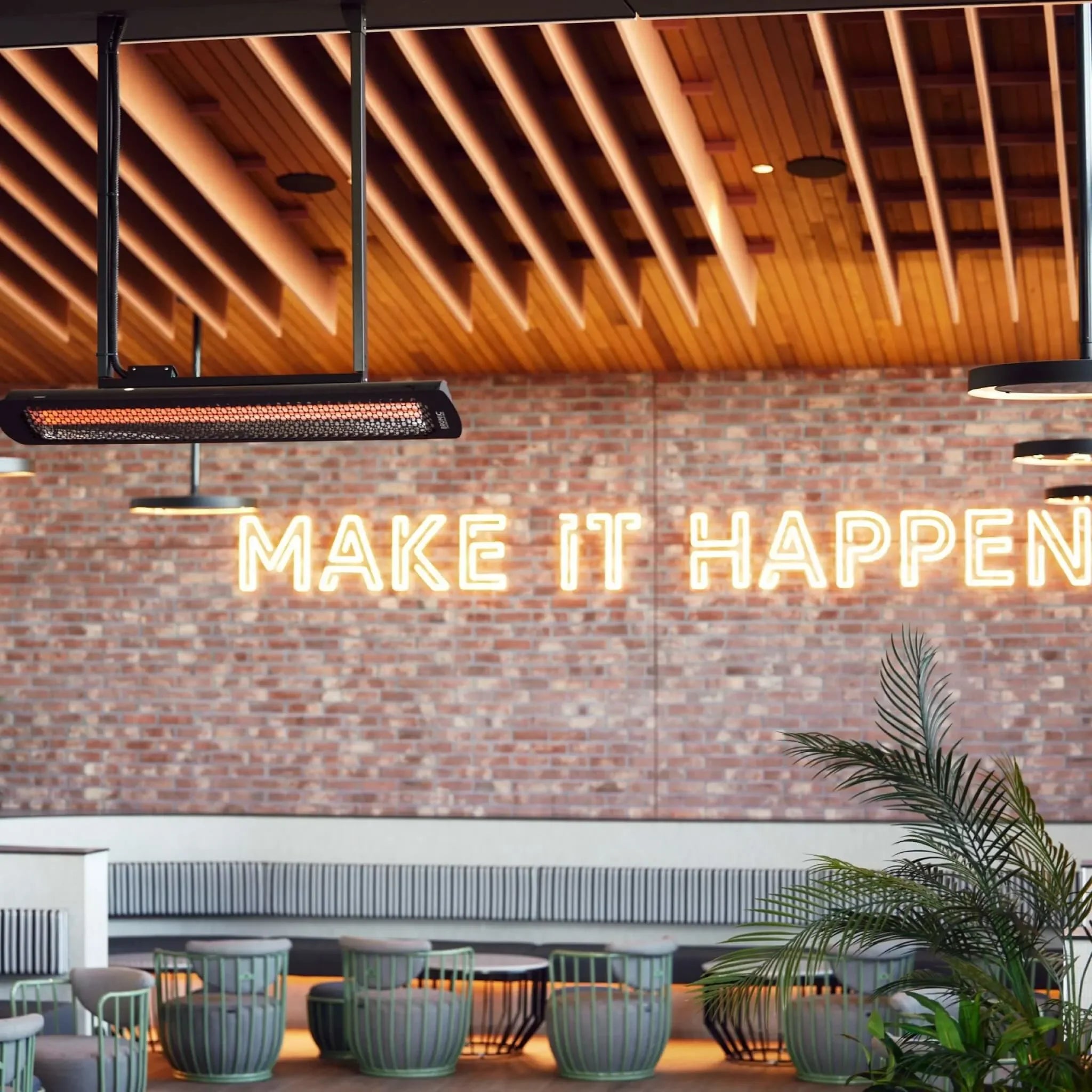Contemporary restaurant interior featuring Bromic Tungsten Electric Smart-Heat heaters ceiling-mounted above a “MAKE IT HAPPEN†neon sign, blending warmth and industrial style for comfortable dining.
