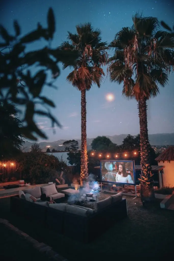 an outdoor movie theater in a backyard - bringing a traditionally indoor space outside. Palm trees sit alongside the screen as the moon rises in the background, comfy outdoor couches situated in front of the movie, string lights for ambiance.