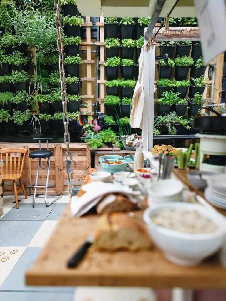 A lovely outdoor kitchen in the daytime; patio bar stools sit at a counter with a spread of food. A outdoor prep station features next to the grill. A vertical garden wall keeps everything feeling more contained.