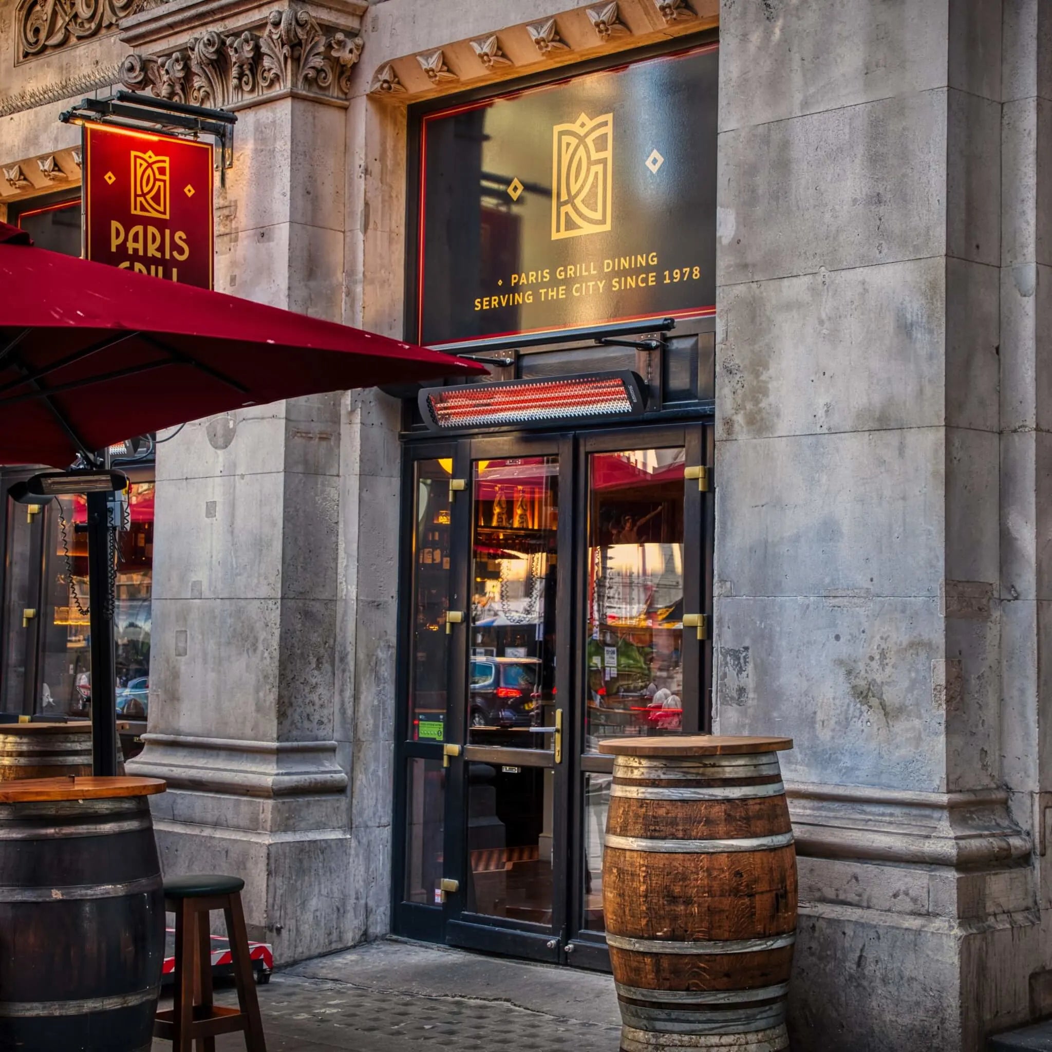 Classic Paris Grill dining entrance in London featuring a Bromic Tungsten Electric Smart-Heat heater above the doorway, providing warmth for guests seated at outdoor barrel tables.